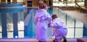 Brother and sister holding hands wearing bathrobes while standing on poolside in hotel